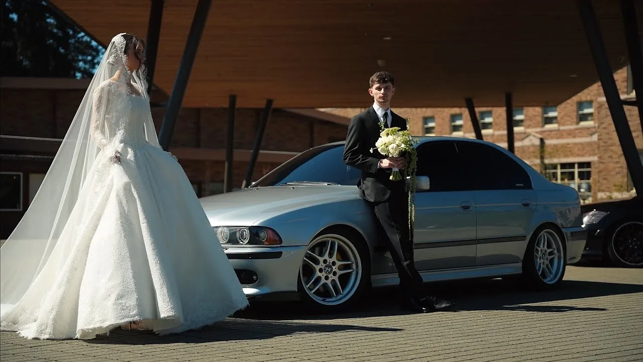 Bride in a long veil and groom with a bouquet beside a silver BMW sedan