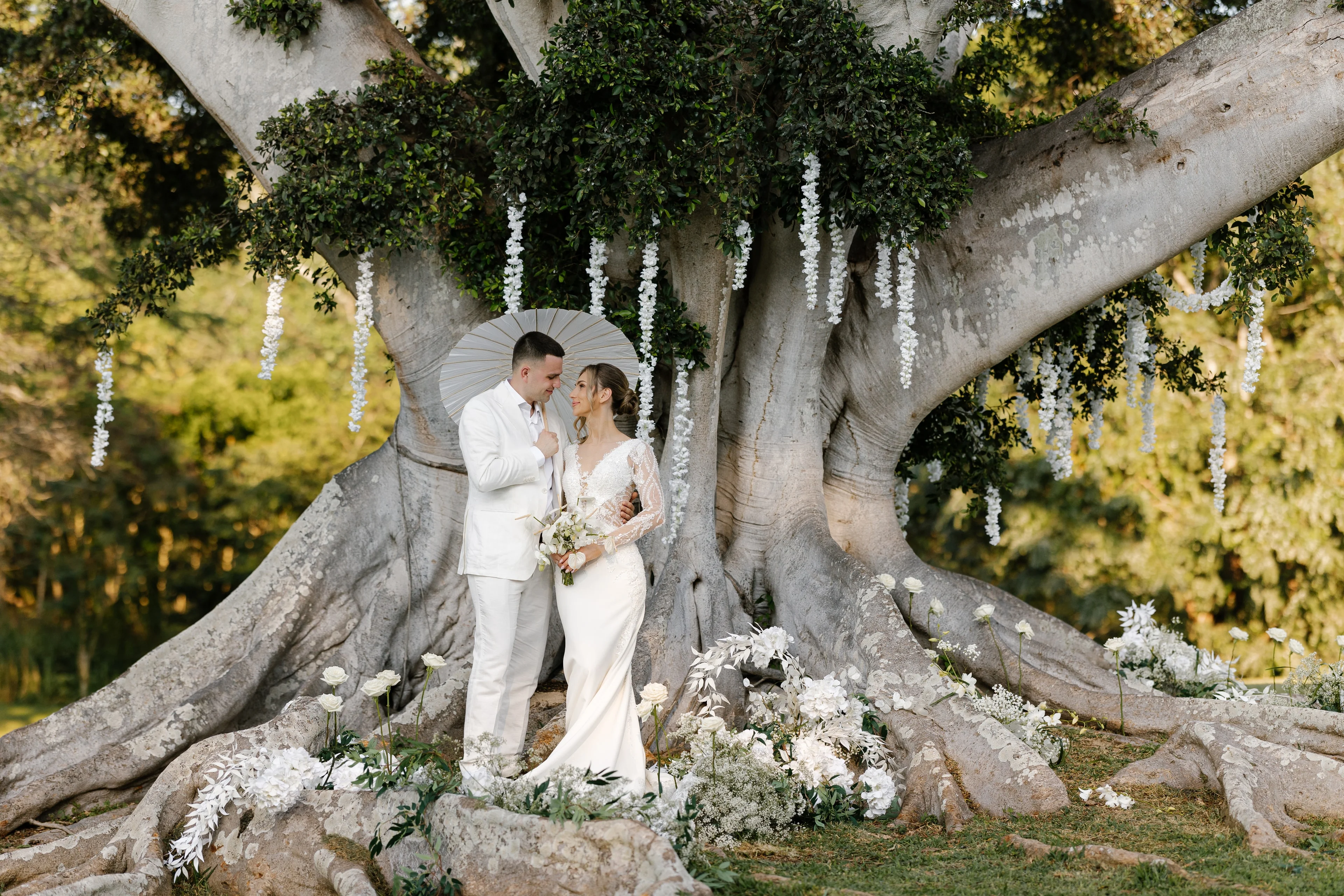 Bride and groom in white under a paper parasol beneath a giant fig tree hung with white flowers