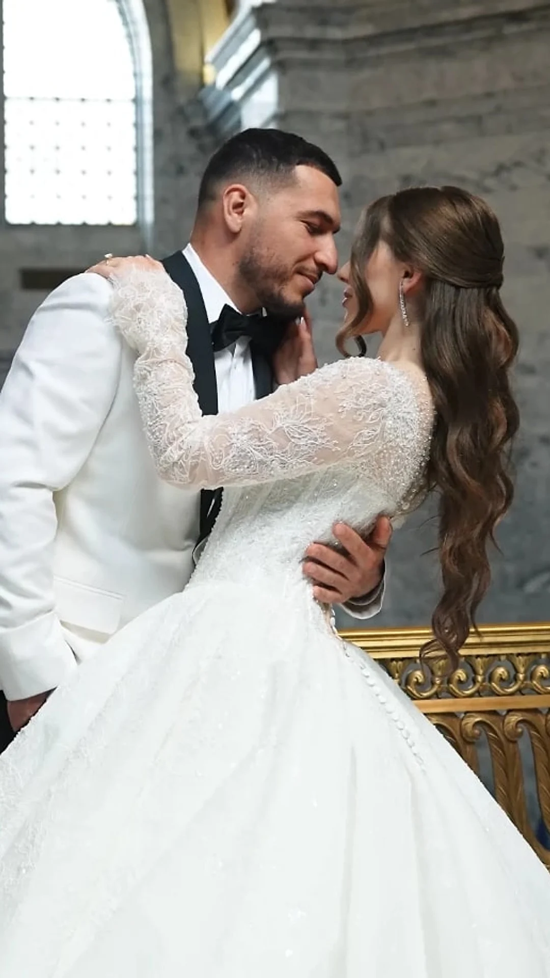 Groom in a white tuxedo and his bride by a gilded railing in a marble hall